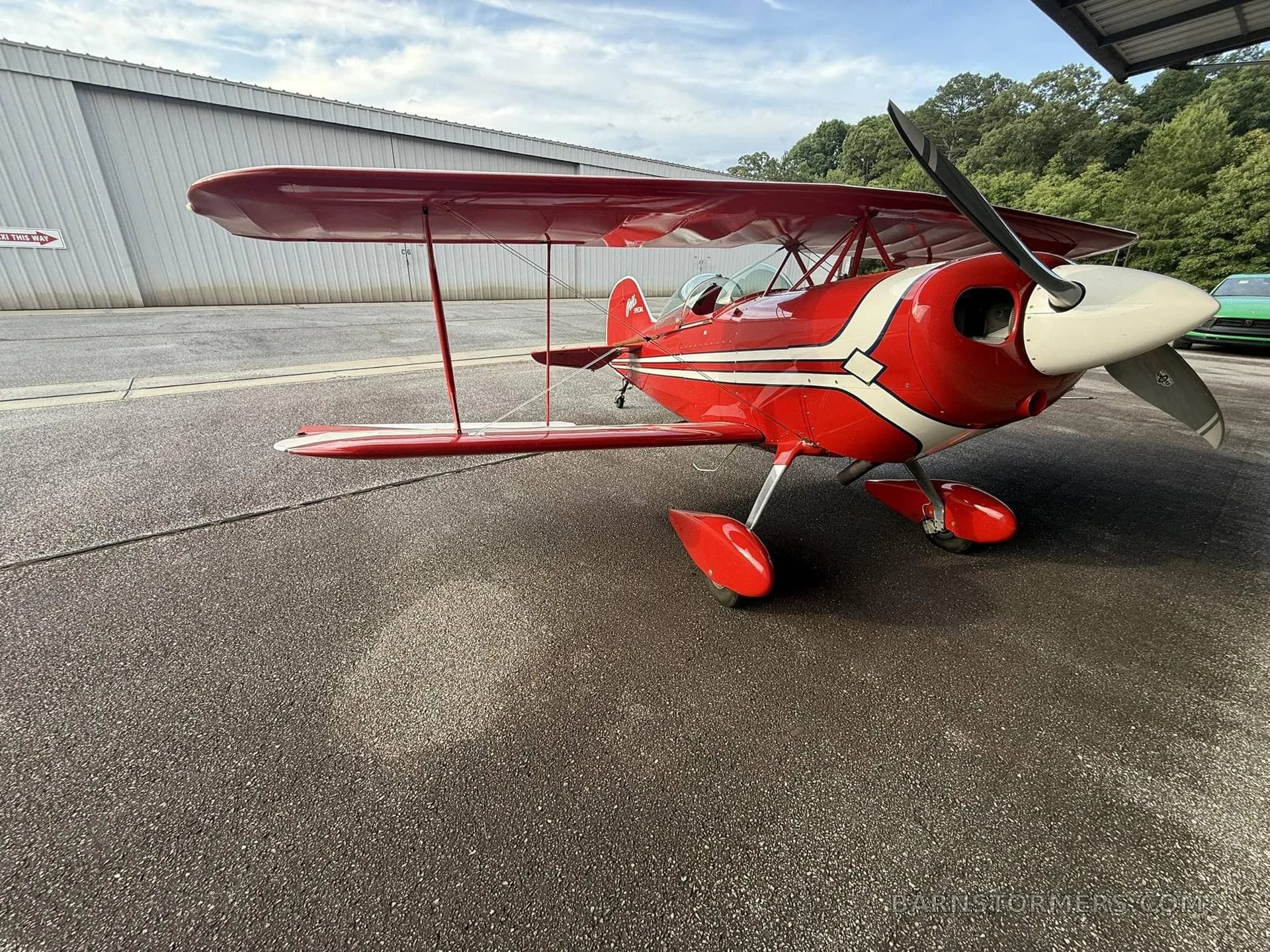A red and white Pitts Special biplane is parked on an asphalt tarmac with a hangar and trees in the background.