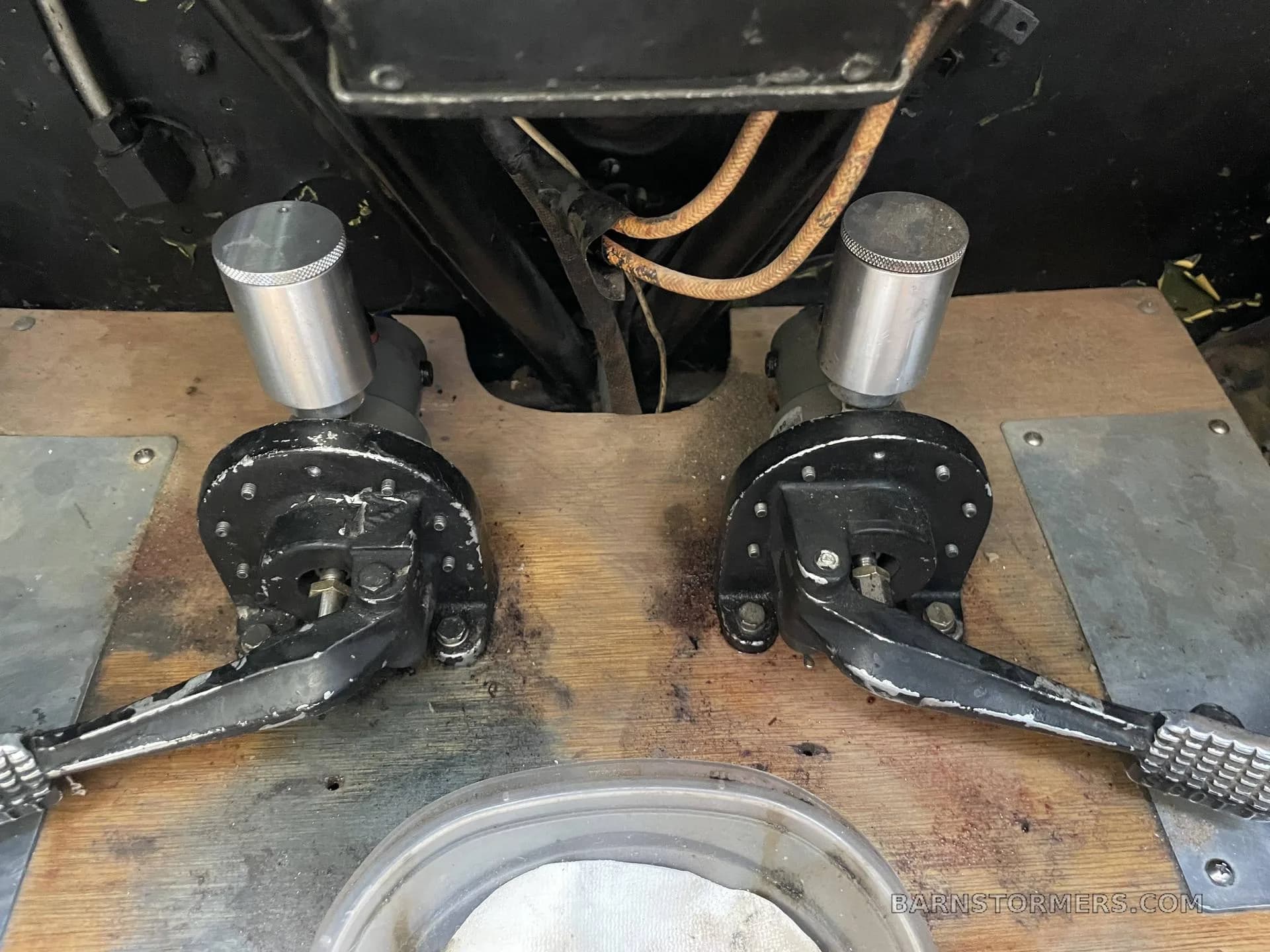 A close-up view of two black rudder pedals and their silver cylinders on a wooden floor in an aircraft cockpit.