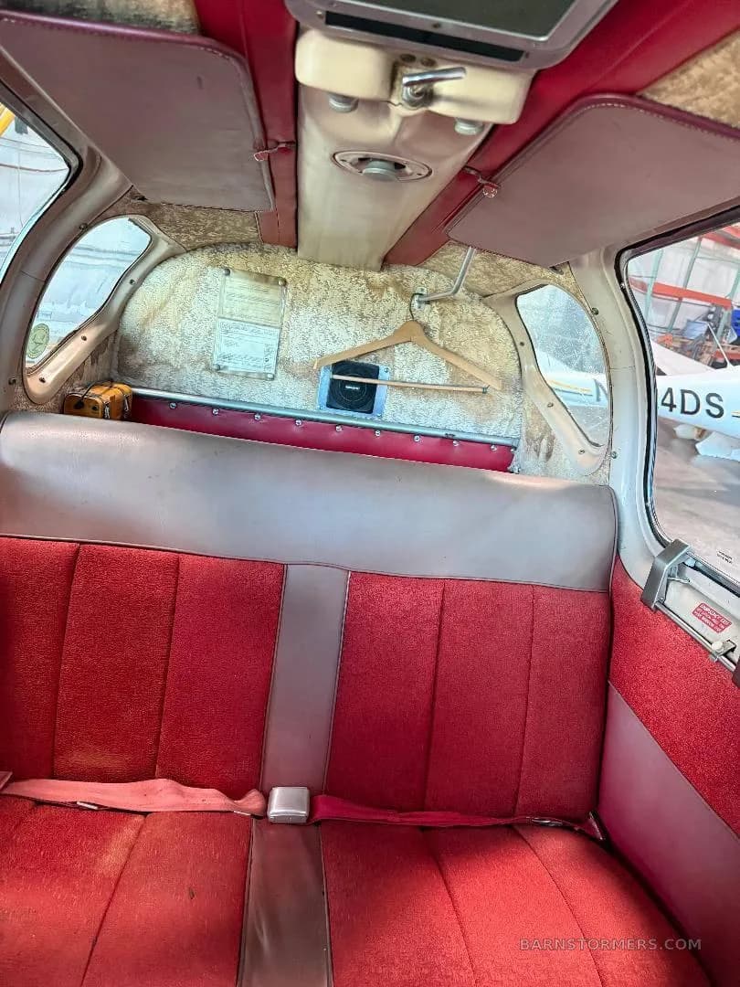 The interior cabin of an aircraft shows red fabric rear seats, a light brown headliner, and a wooden clothes hanger.
