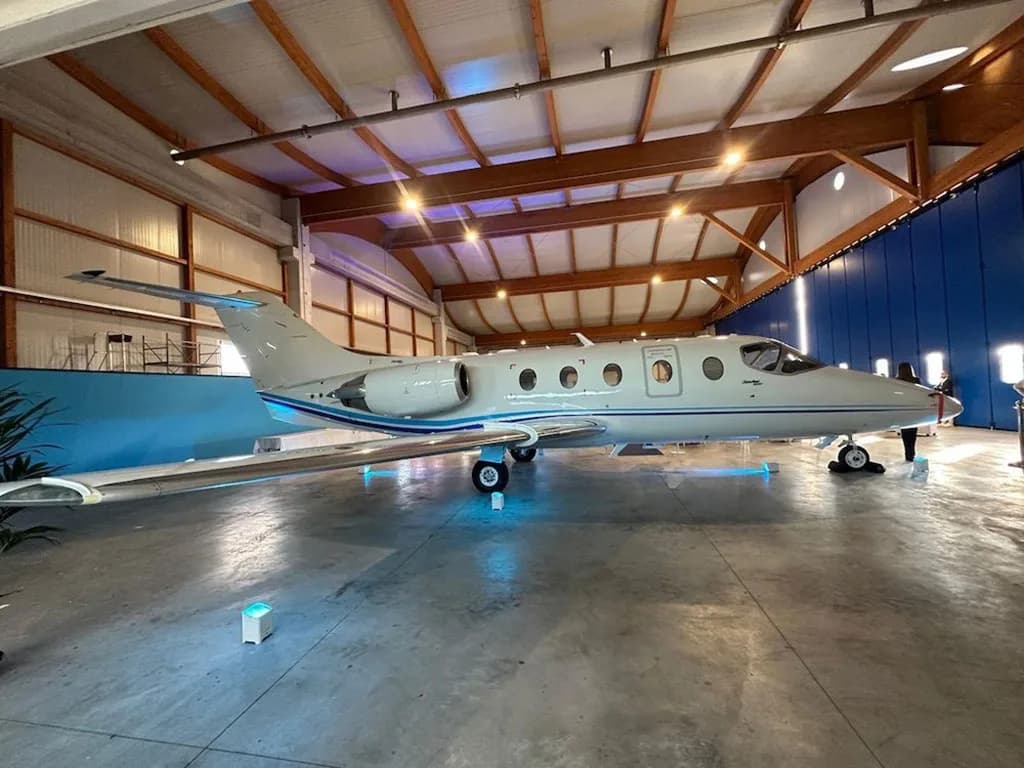 A white business jet with blue stripes is parked in a hangar, viewed from the side.