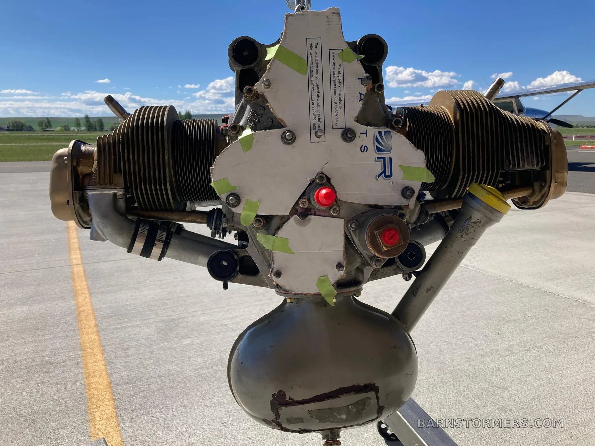 The front view of an aircraft engine with a covered propeller hub, suspended on an airfield tarmac.