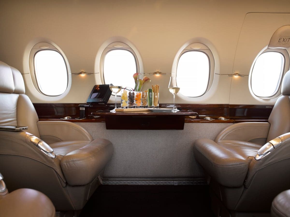 Two tan leather seats face each other in a private jet cabin, with a table holding drinks and snacks.