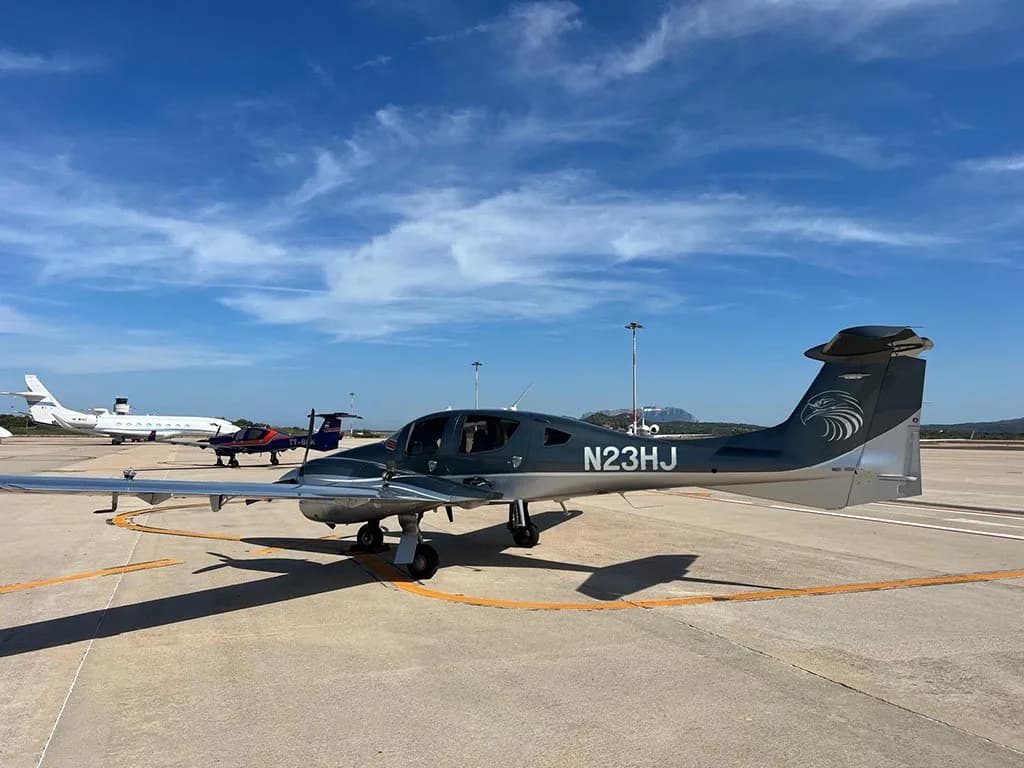 A side view of a dark grey Diamond DA62 aircraft with registration N23HJ and an eagle graphic on the tail, parked on an airport tarmac.