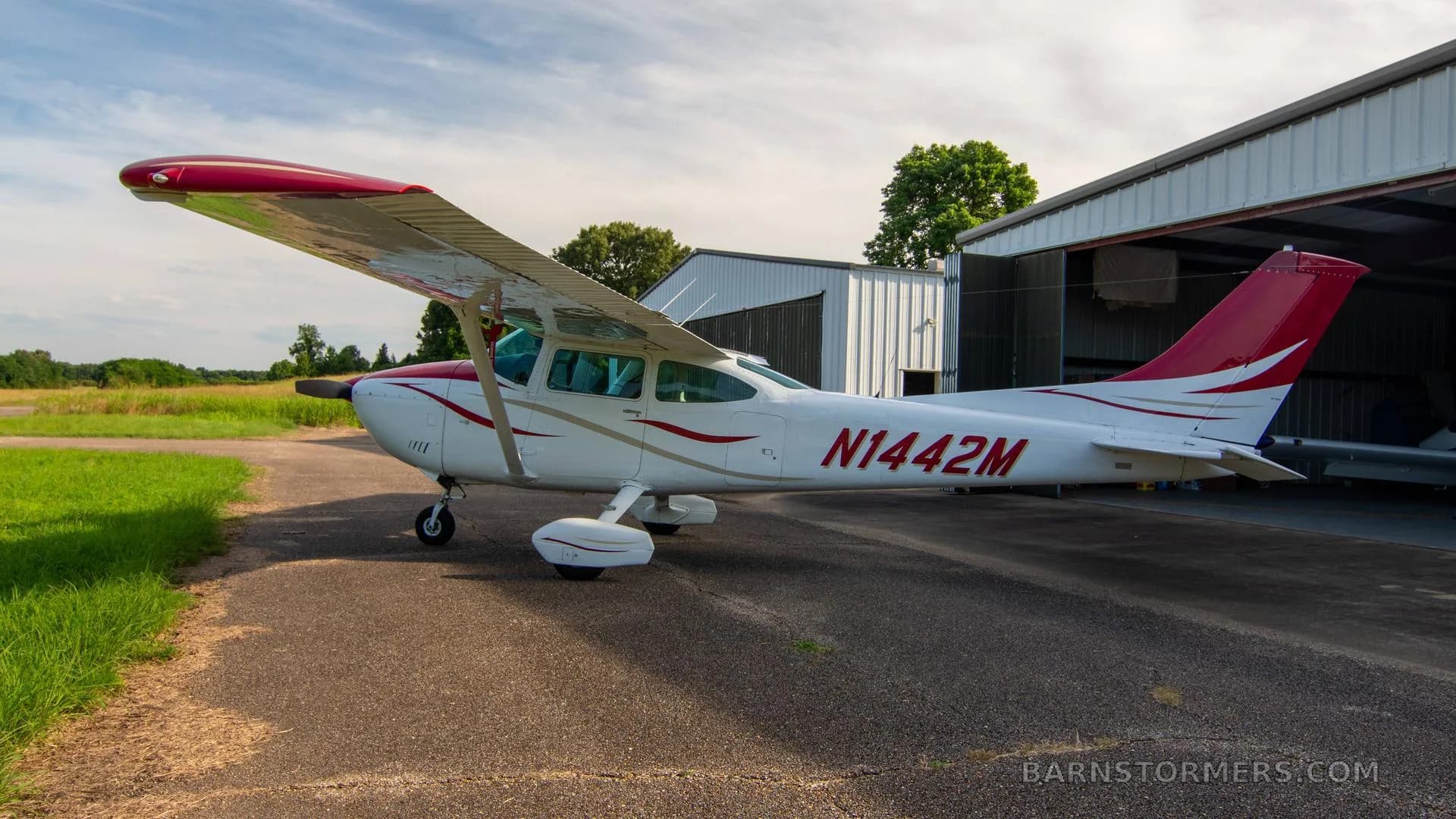 A white single-engine Cessna aircraft with red and gold stripes is parked on an asphalt ramp, viewed from the front-right.