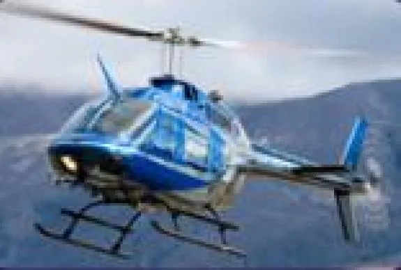 Blue and silver Bell 206B helicopter flying in front of a mountain.