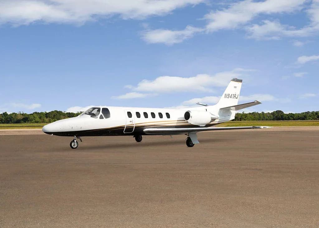 A white and black Cessna Citation business jet N941RJ with gold stripes is parked on a tarmac under a partly cloudy sky, viewed from the front-left.