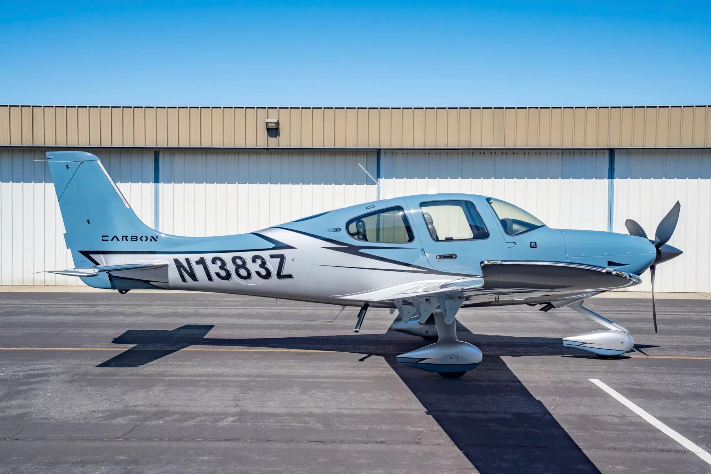 A light blue and silver Cirrus SR22T aircraft, N1383Z, parked on an tarmac in front of a hangar.