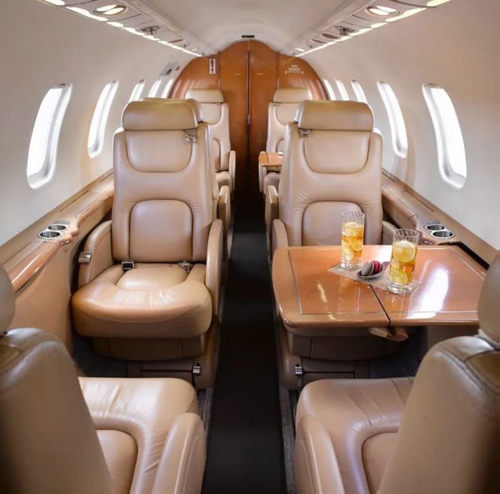The aircraft cabin features tan leather seats, wooden tables with drinks, and overhead lighting.