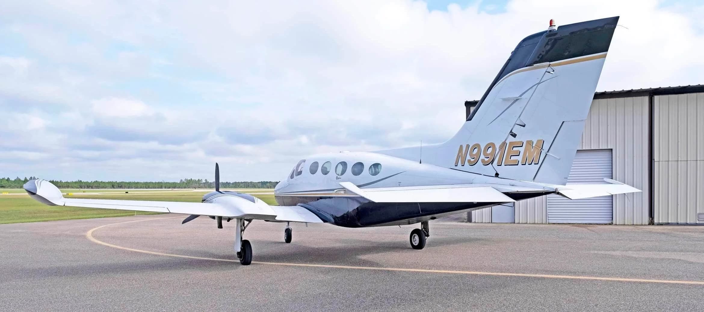 A rear-quarter view of the white and dark blue Cessna 414 Chancellor, registration N991EM, parked on an airfield near a hangar.