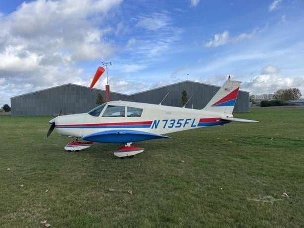 A Piper PA-28-181 Archer II aircraft, registration N735FL, is parked on a grassy field with a windsock nearby.