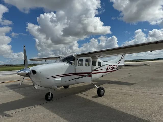 A three-quarter front view of a white Cessna 206 with red and grey stripes, showing the tail number N7567K, parked on an airfield.