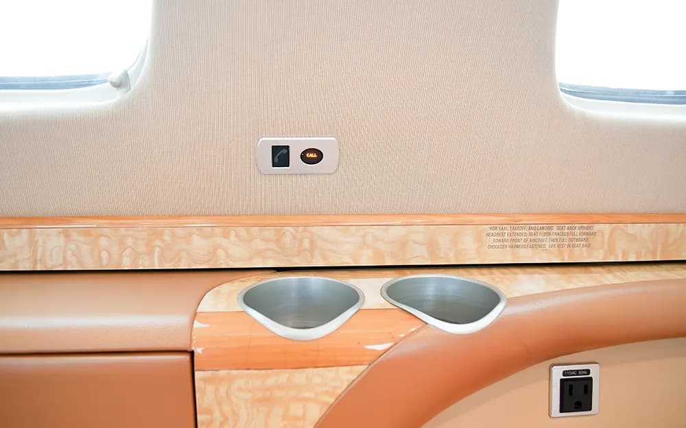 A close-up of an aircraft cabin wall with a call button, power outlet, and two cup holders in a wood-paneled console.