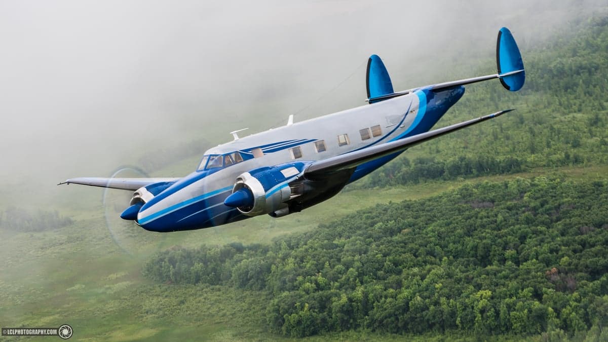 A blue and silver Beechcraft Model 18 flying over a green, forested area.