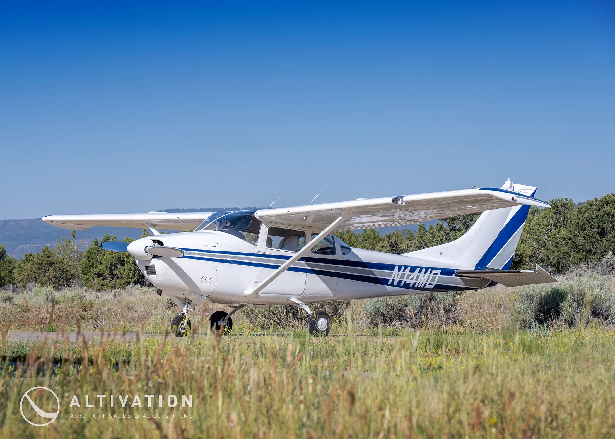 A white Cessna 182 with blue and grey stripes, registration N74MD, parked on a paved surface next to grassy terrain under a clear blue sky.