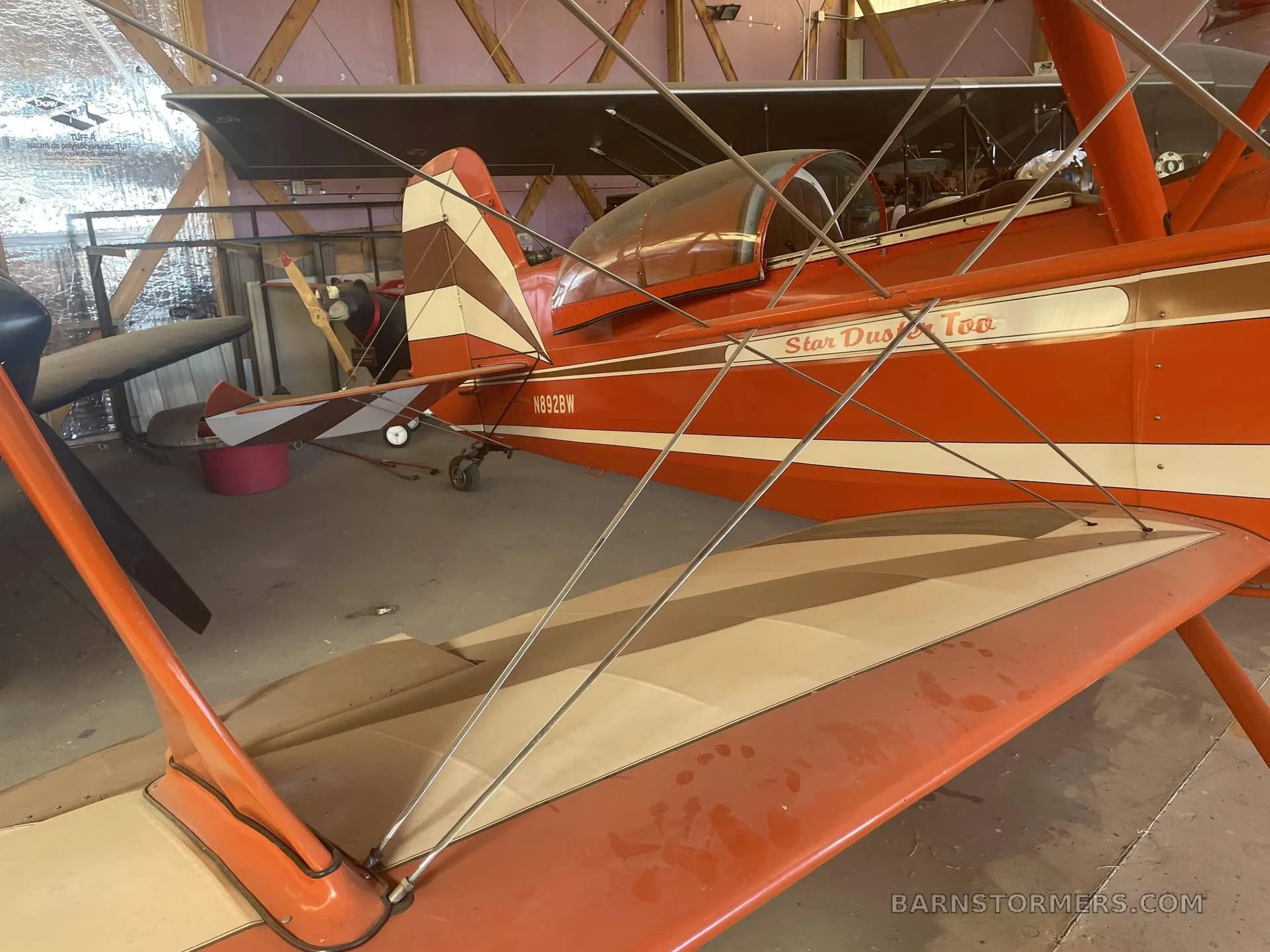 A rear-side view of an orange and white Star Duster Too biplane with its tail section visible, parked in a hangar.