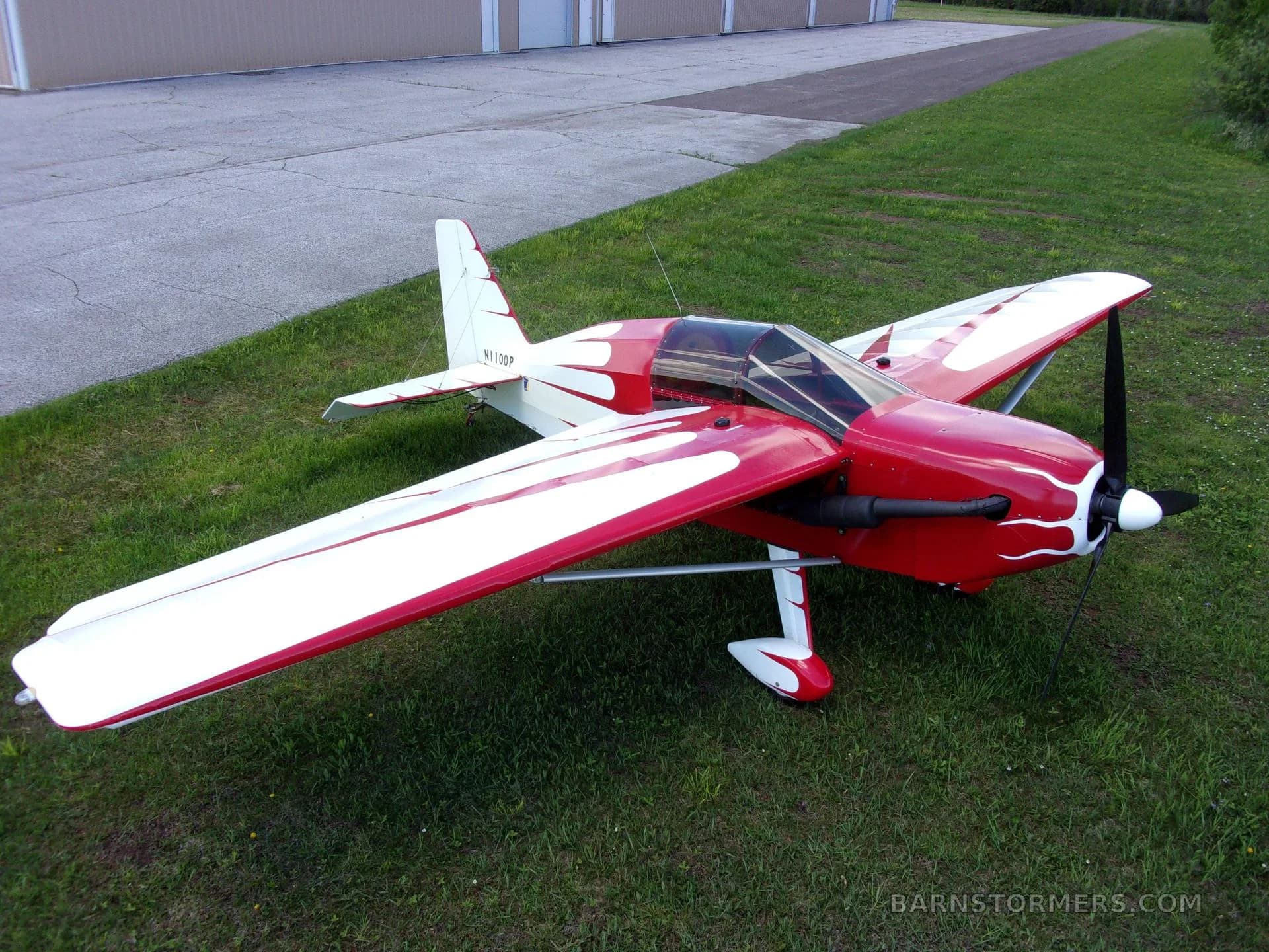 A red and white single-engine aircraft with a flame-like paint scheme, parked on grass next to a hangar.