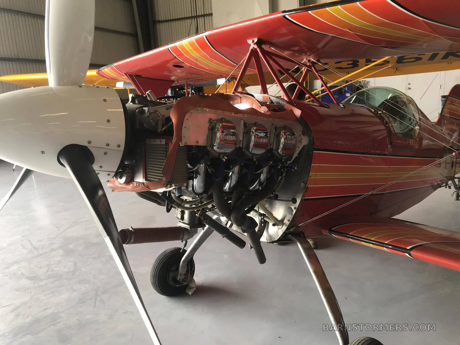 A close-up view of the red biplane's engine, with the cowling removed, showing the cylinders and exhaust in a hangar.