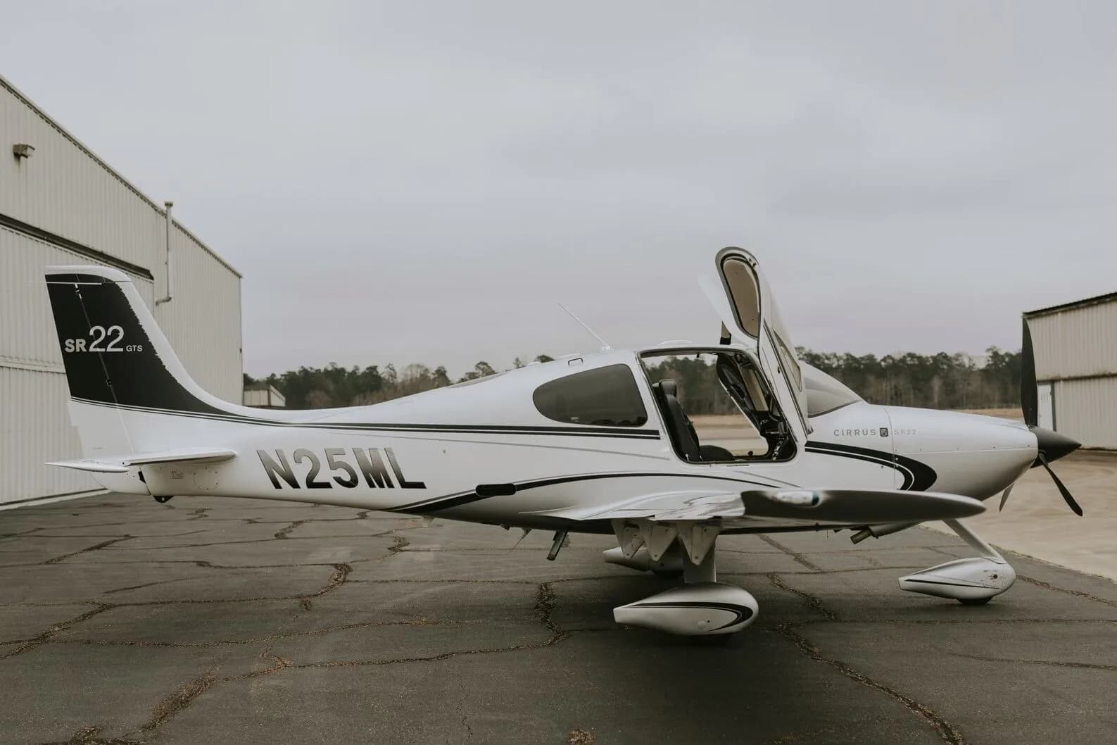 A silver Cirrus SR22 GTS aircraft with its gull-wing doors open, parked on an asphalt ramp with a hangar in the background.