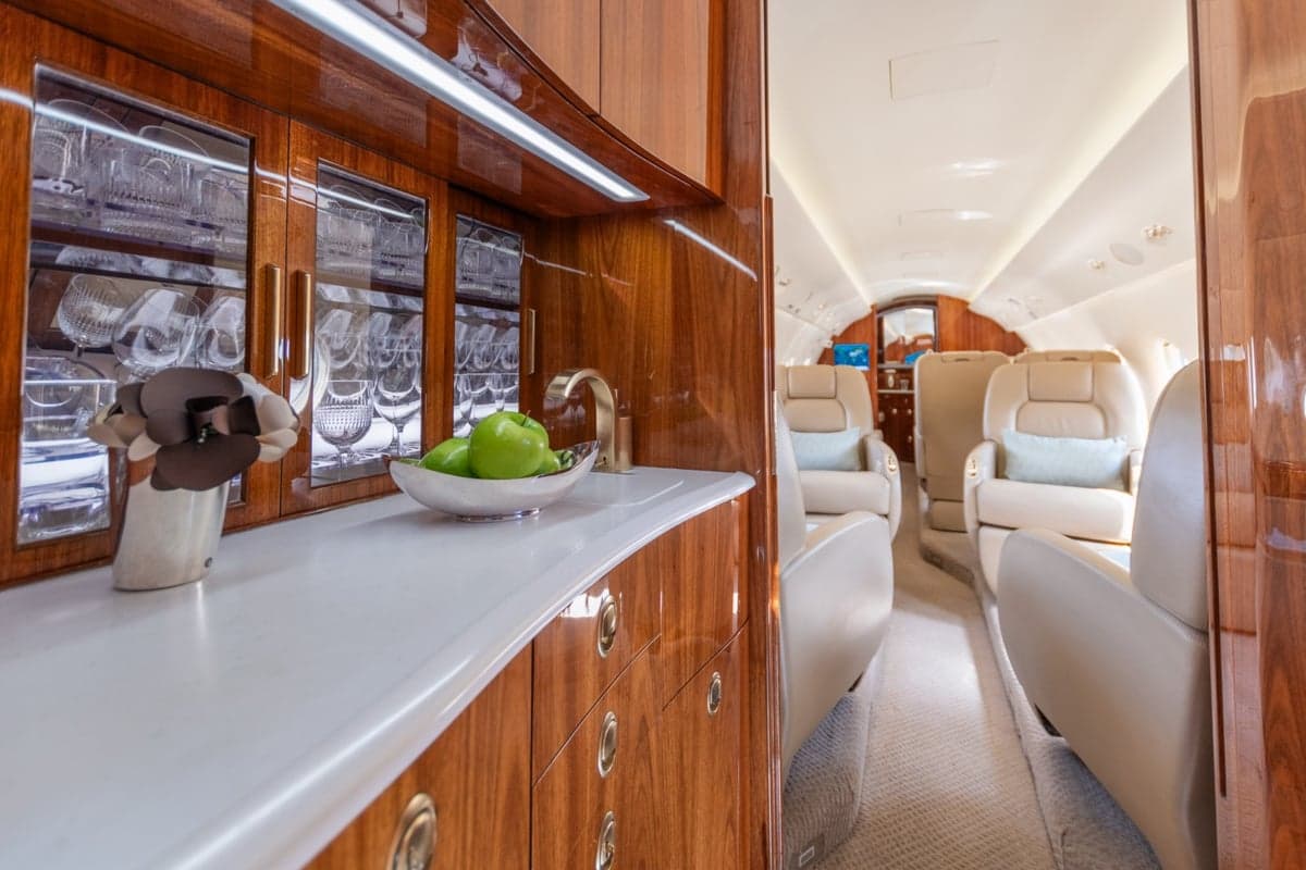 A well-appointed aircraft galley with polished wooden cabinetry, a white countertop, glassware, and a bowl of green apples.