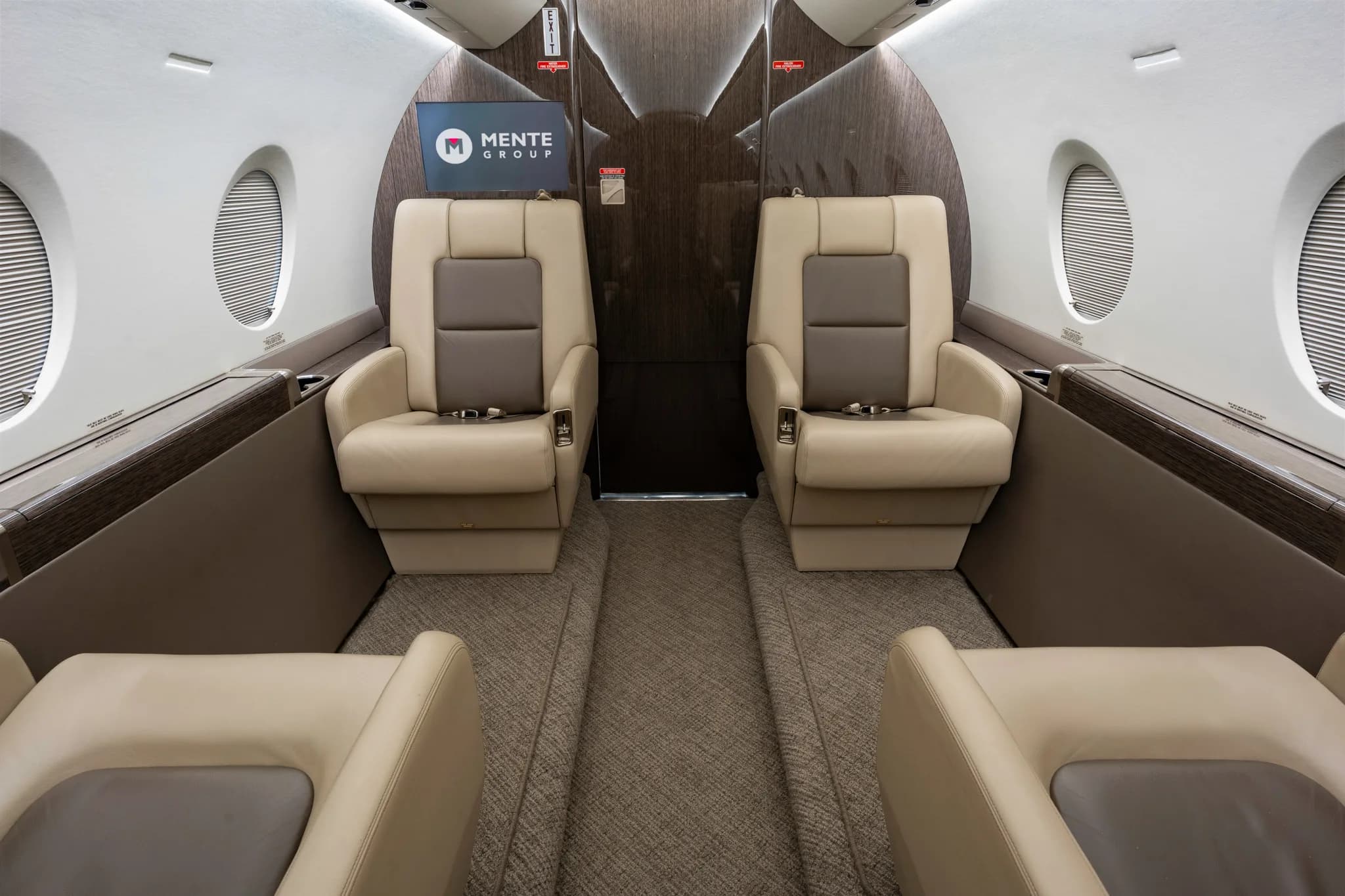 The interior of a private jet cabin, showing an aisle between two rows of comfortable beige armchairs and a screen displaying a logo at the rear.