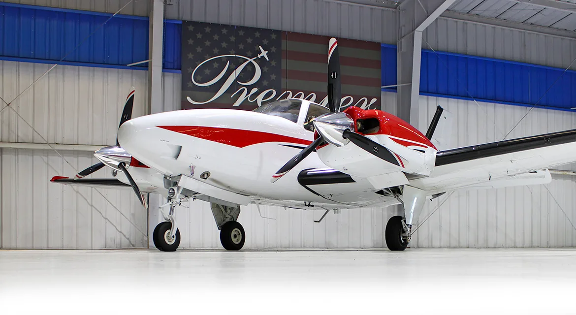 A front-quarter view of a white and red twin-engine propeller aircraft, a Beechcraft Baron, in a hangar.