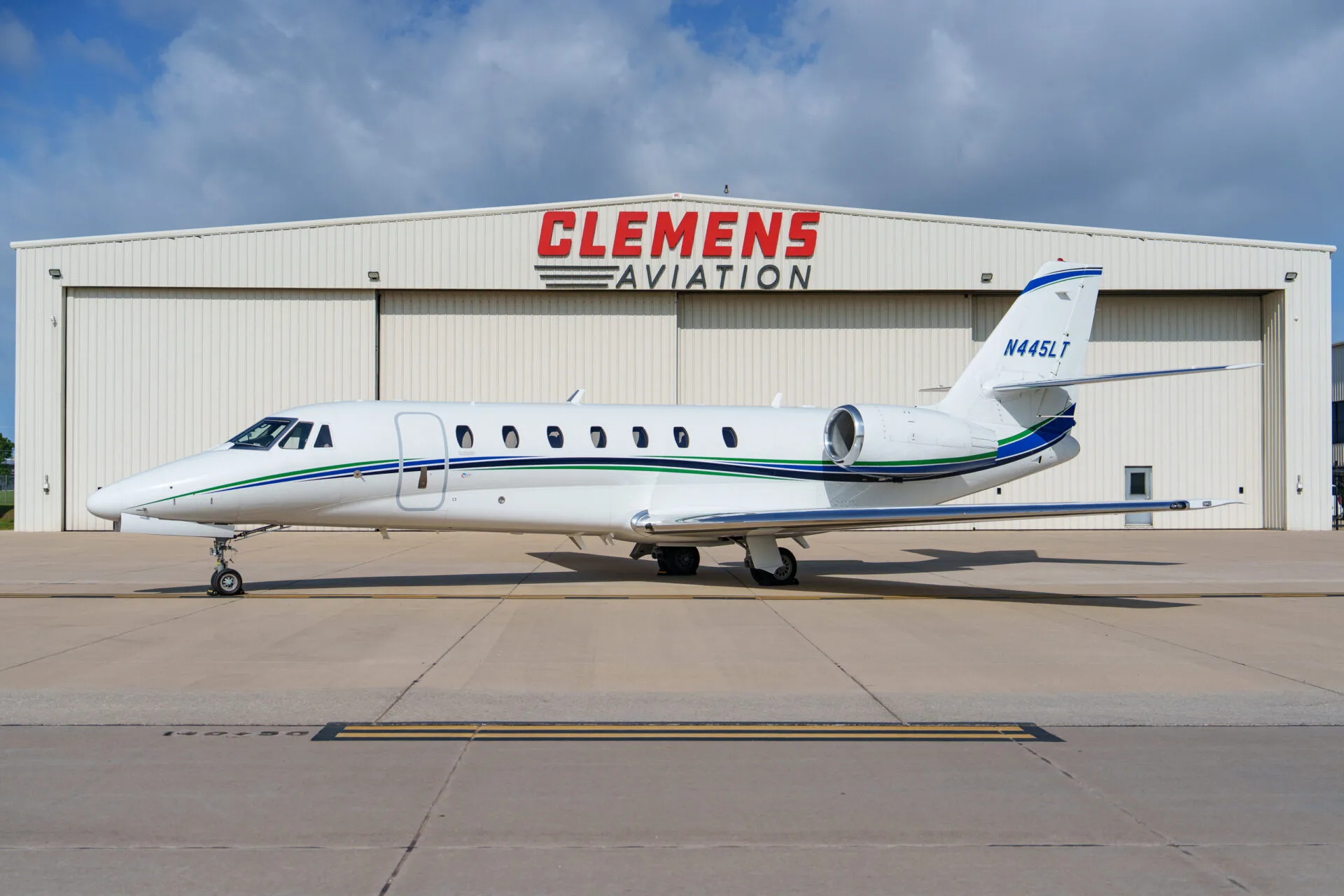 A white Cessna Citation business jet with green and blue stripes parked in front of a "Clemens Aviation" hangar.