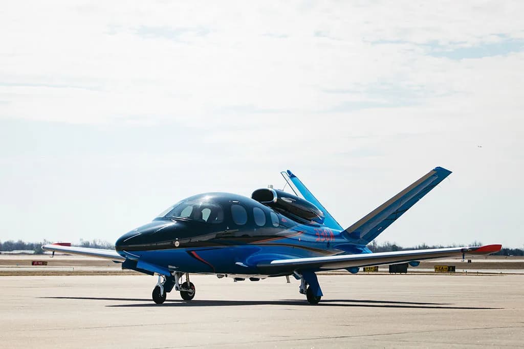 A black and blue Cirrus Vision Jet (SF50) with red accents parked on an airfield under a partly cloudy sky.