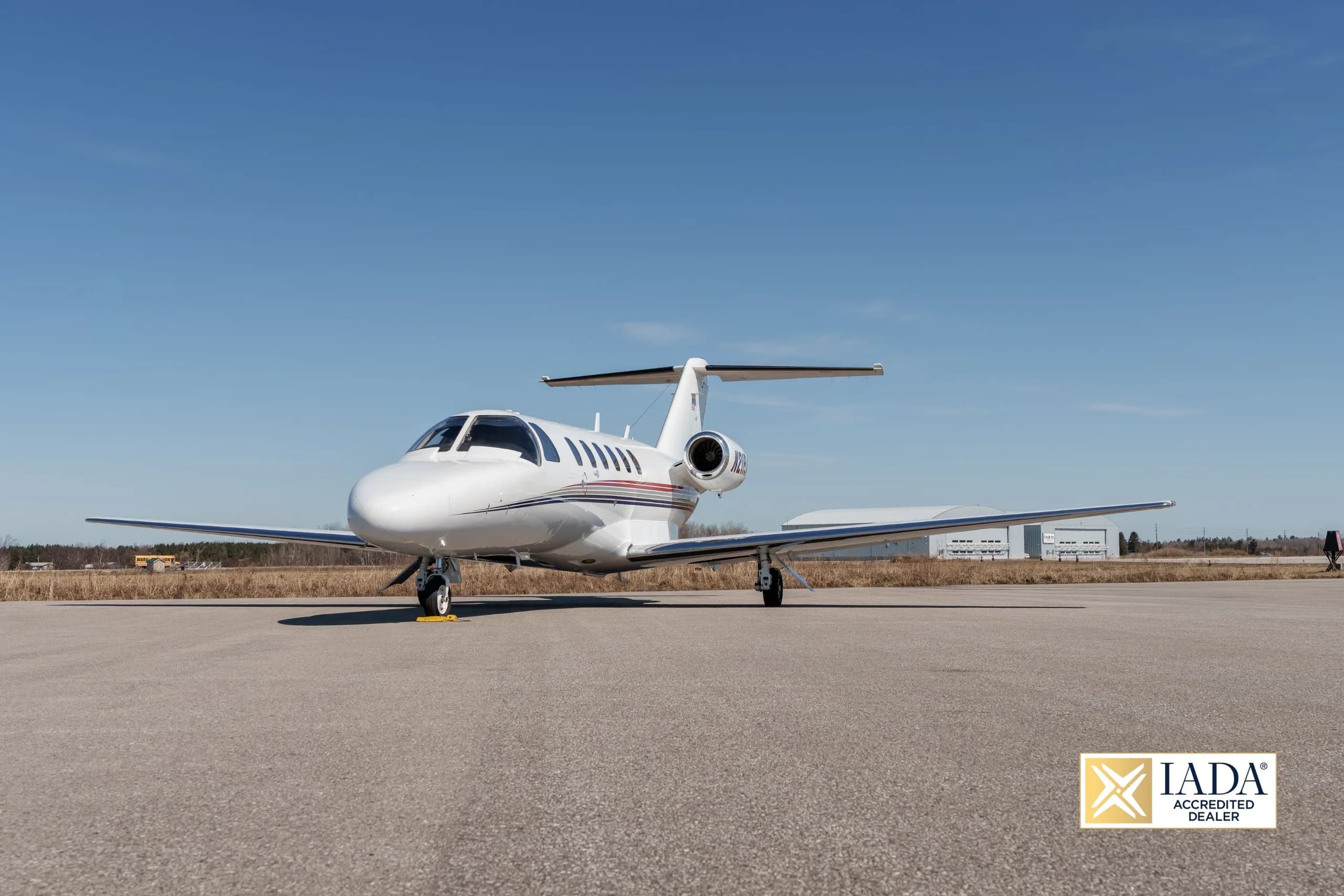 A white Cessna Citation business jet (N21RA) with red and blue stripes parked on an airfield, viewed from the front.