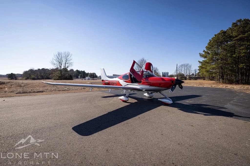 A red and silver Cirrus SR22T aircraft with open doors parked on an asphalt ramp with trees in the background.