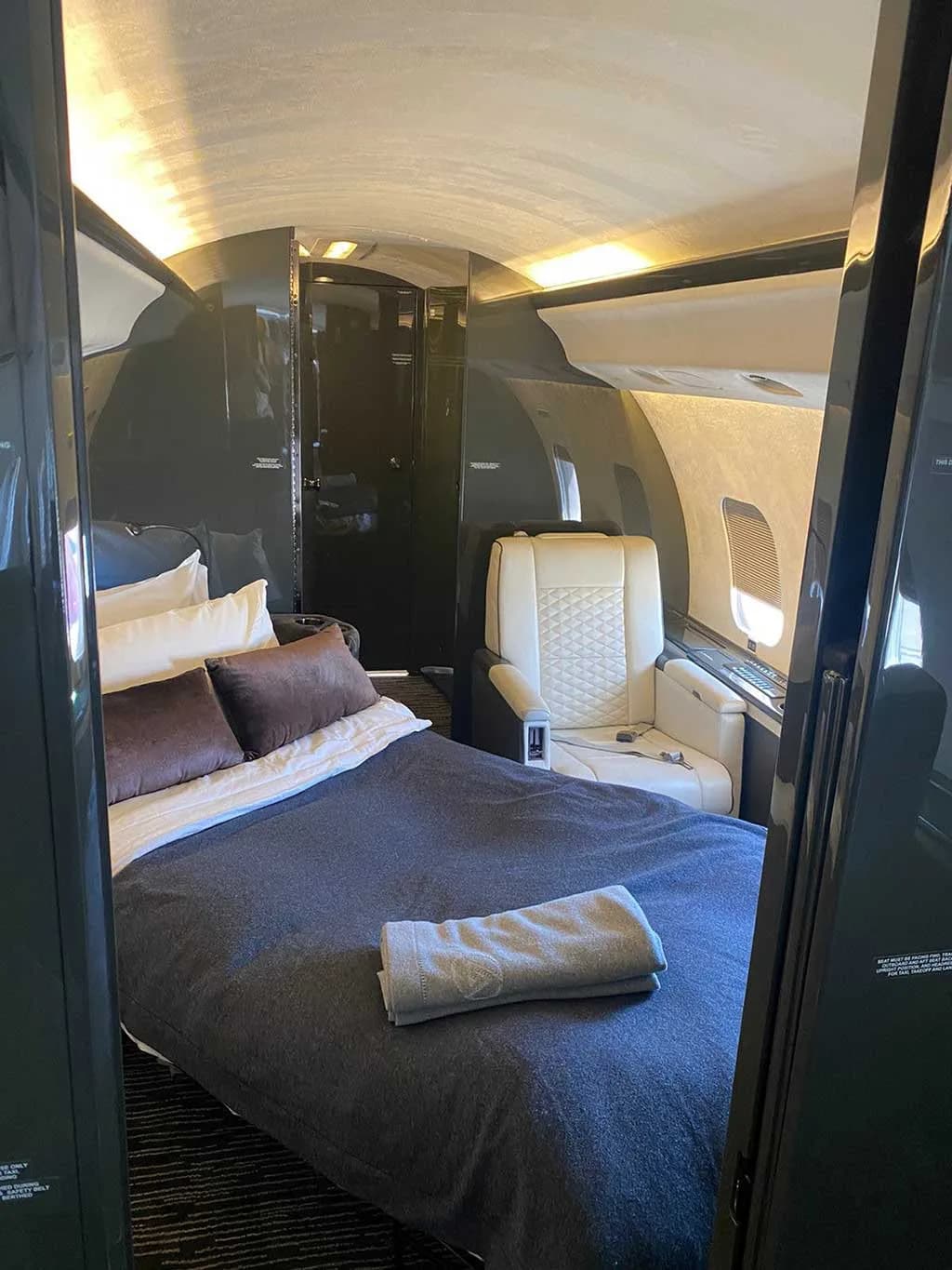 The private jet cabin converted into a bedroom, featuring a dark blue bed with pillows and a folded towel, next to a white quilted seat.