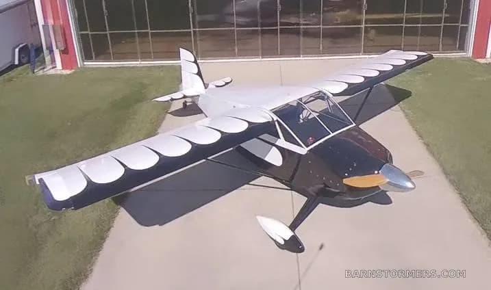 An overhead view of a black and white single-engine aircraft with a wooden propeller parked on concrete in front of a red hangar.