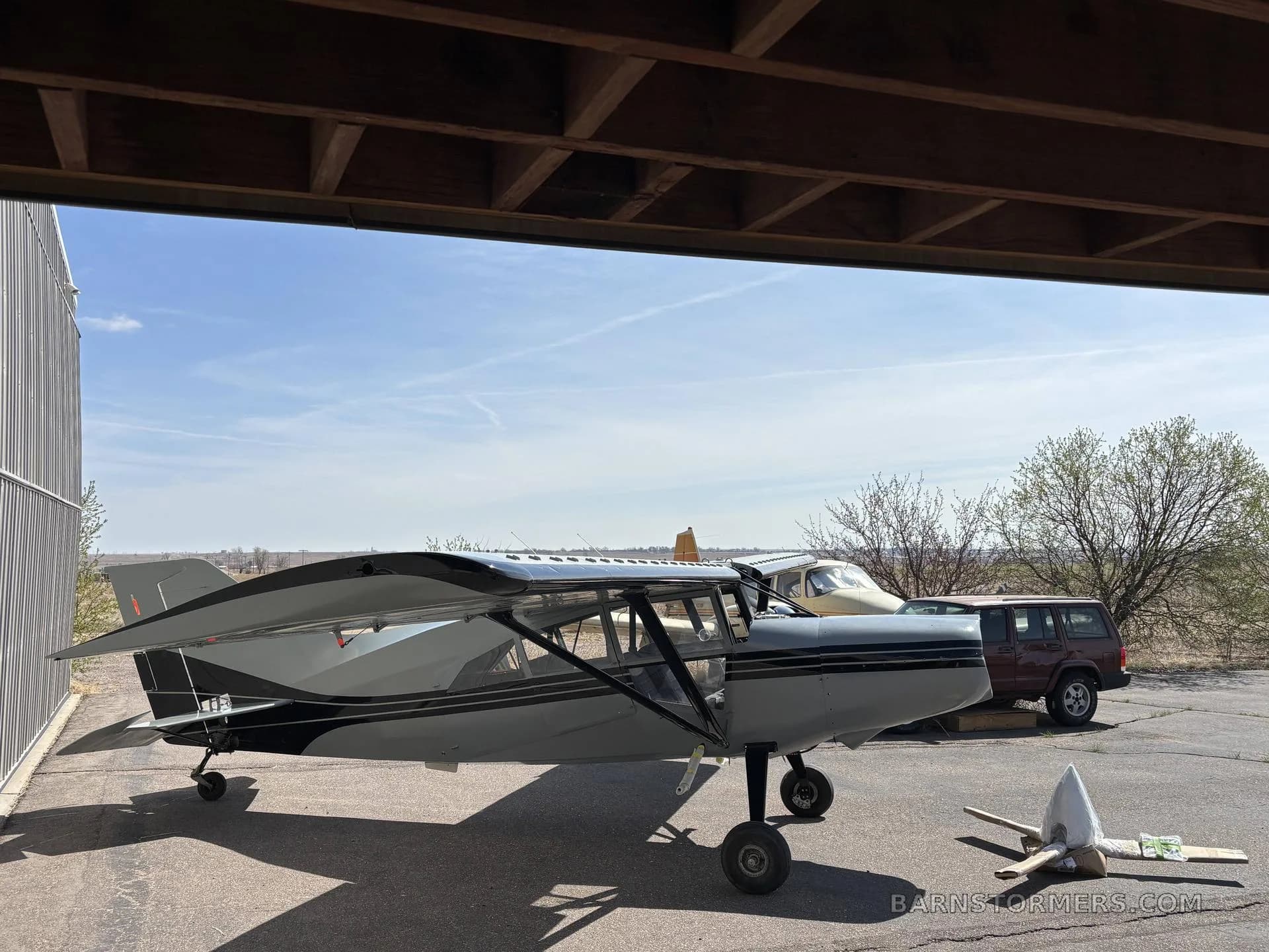 A grey Maule M-7 aircraft with black stripes parked under an overhang, with another aircraft and vehicle in the background.