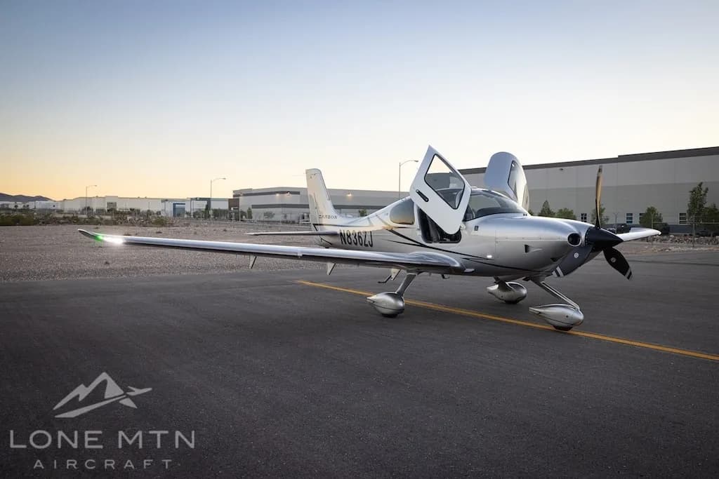 A silver Cirrus SR22T aircraft with registration N836ZJ is parked on an asphalt ramp with its doors open, showing its front-right profile with navigation lights illuminated.