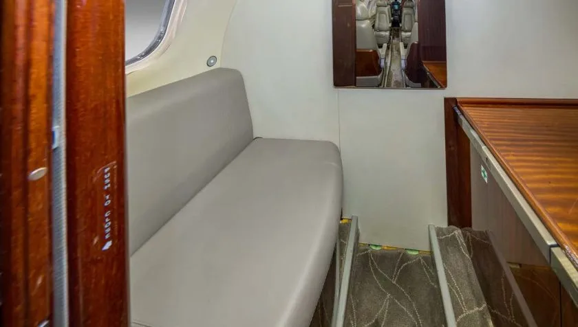 A view inside the cabin of a private jet, showing a light grey bench seat and a wooden cabinet.