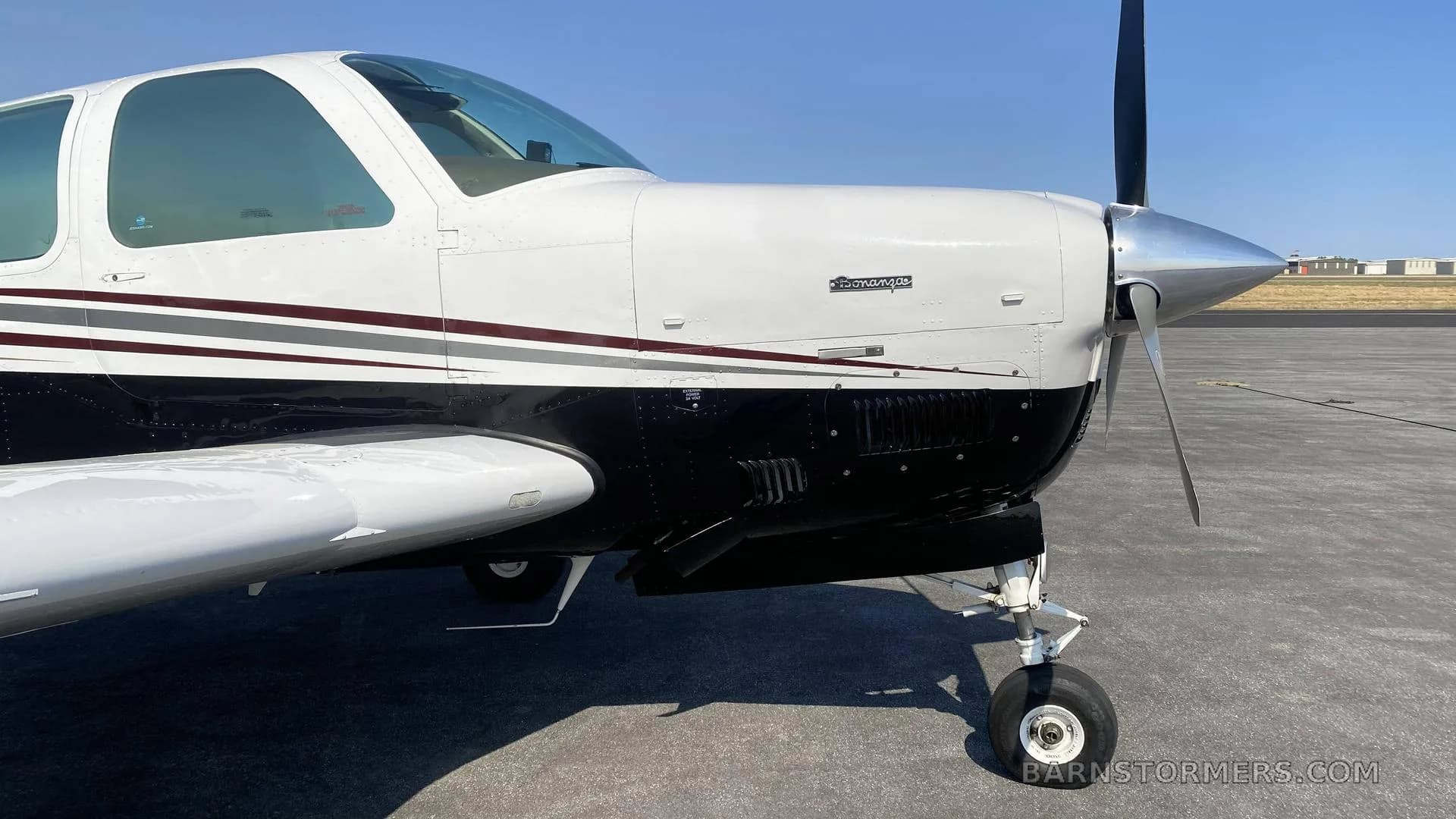 A close-up view of the nose and propeller of a white and black single-engine aircraft, showing the engine cowling and landing gear.