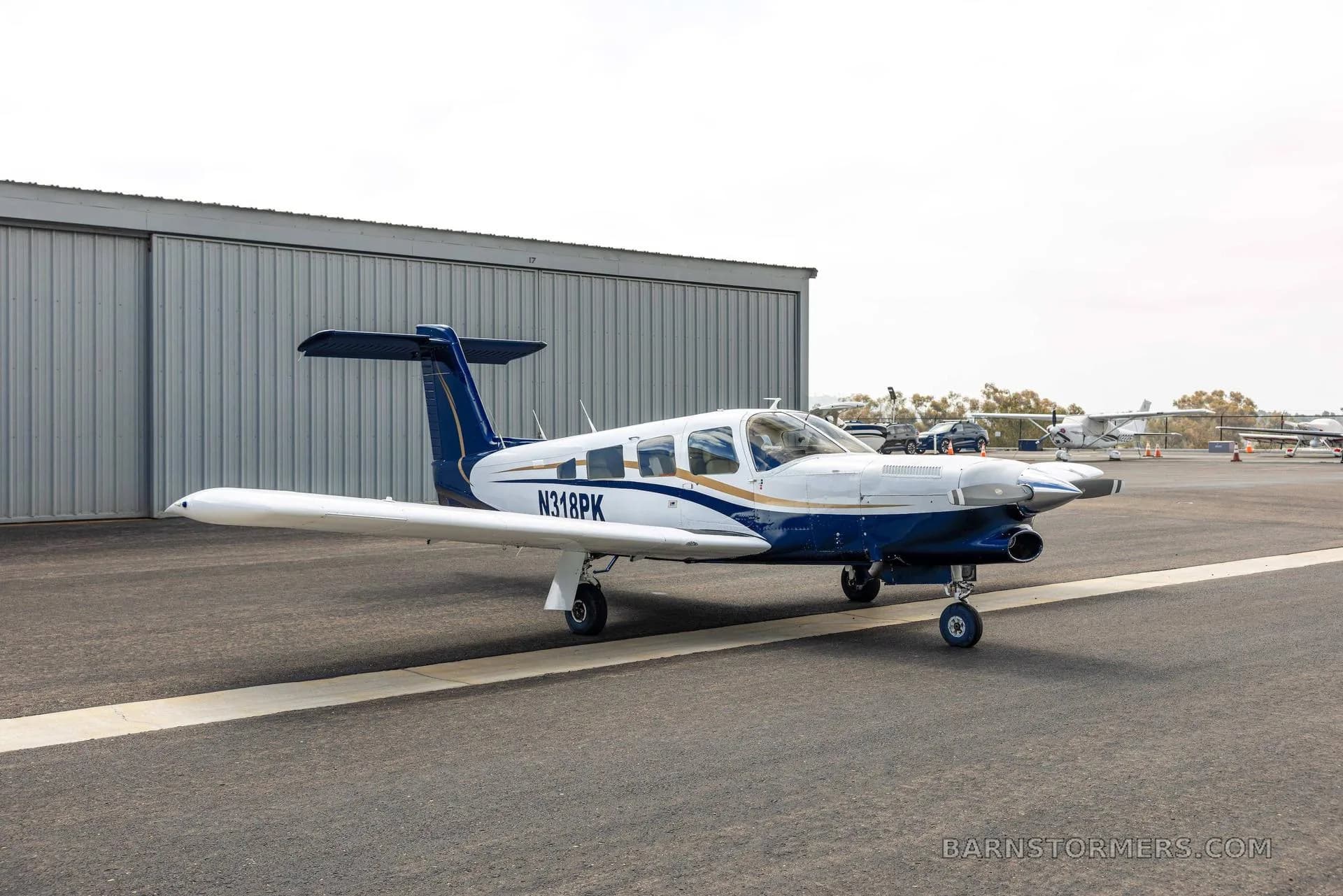 A three-quarter rear view of a white and blue single-engine propeller aircraft with tail number N318PK, parked on an asphalt tarmac.