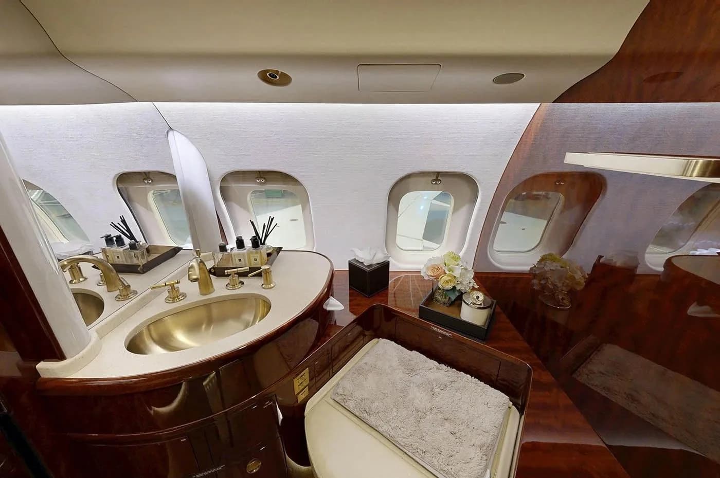 A luxurious aircraft lavatory with a gold-colored sink, dark wood vanity, and a toilet with a grey mat, all illuminated by natural light from the windows.