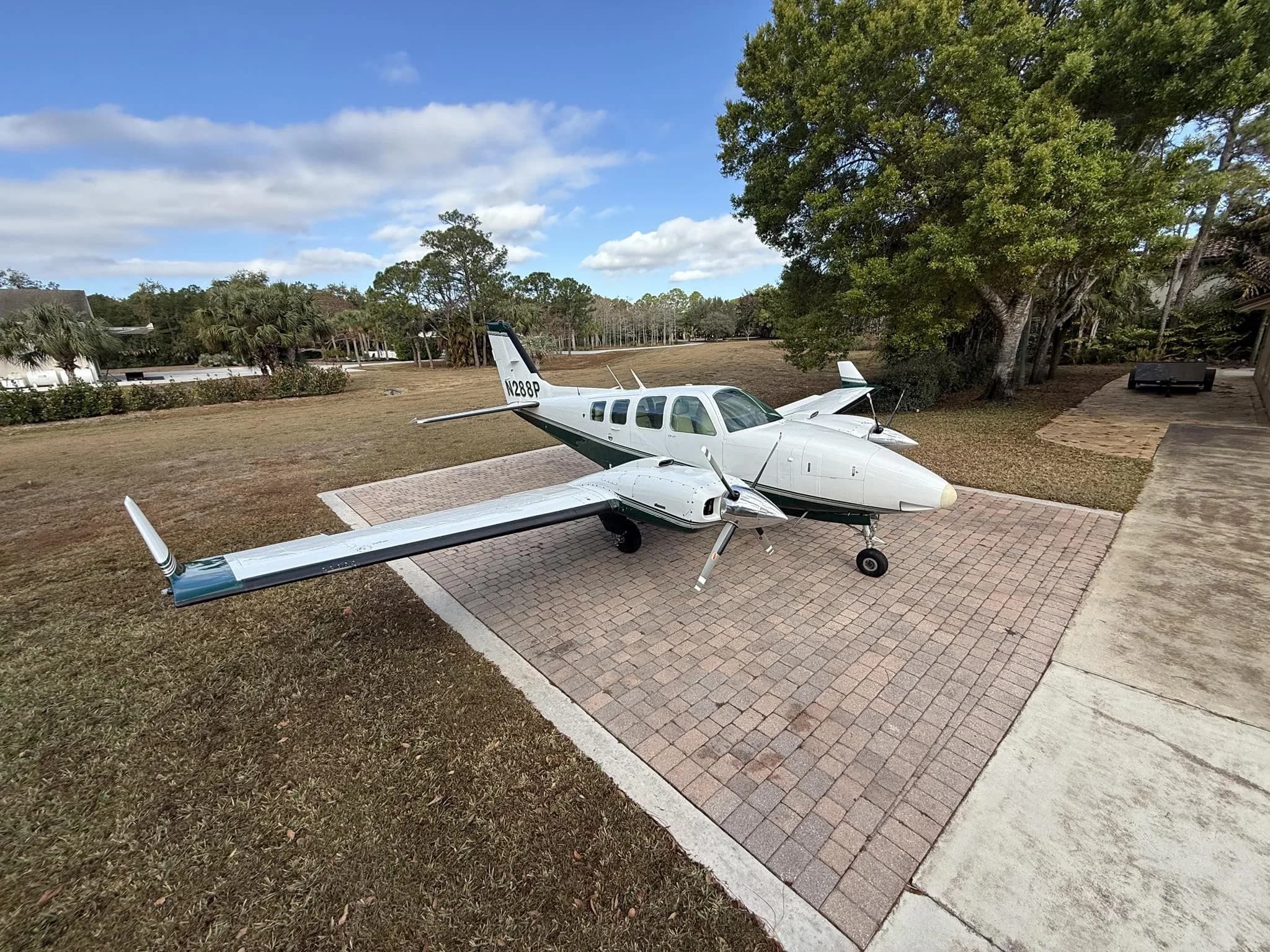 A white and green twin-engine Beechcraft Baron aircraft with tail number N289P parked on a brick-paved area next to a grassy field and trees under a partly cloudy sky.