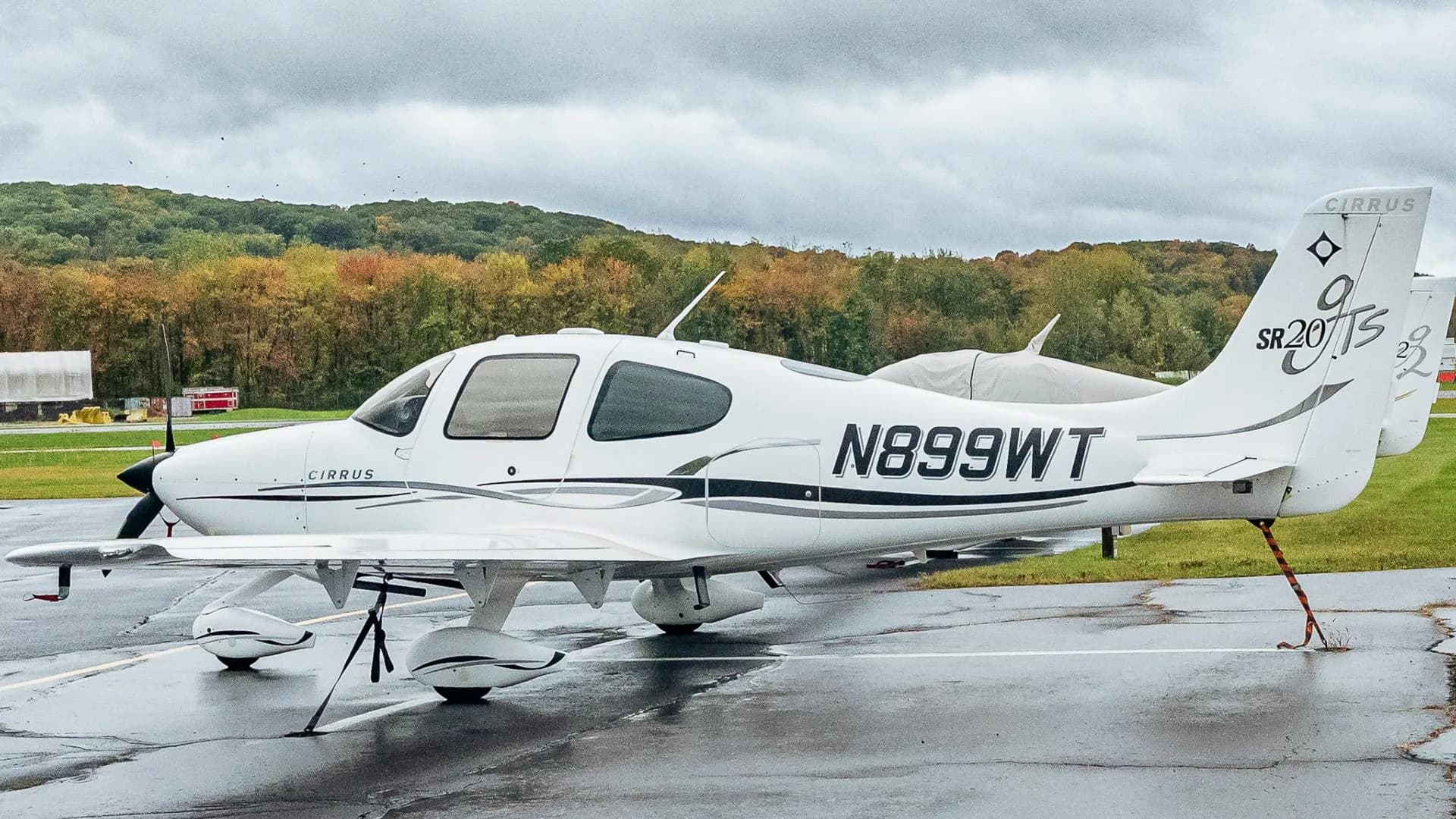 A white Cirrus SR20 aircraft with black and grey accents, registration N899WT, parked on a wet tarmac with autumn trees in the background.