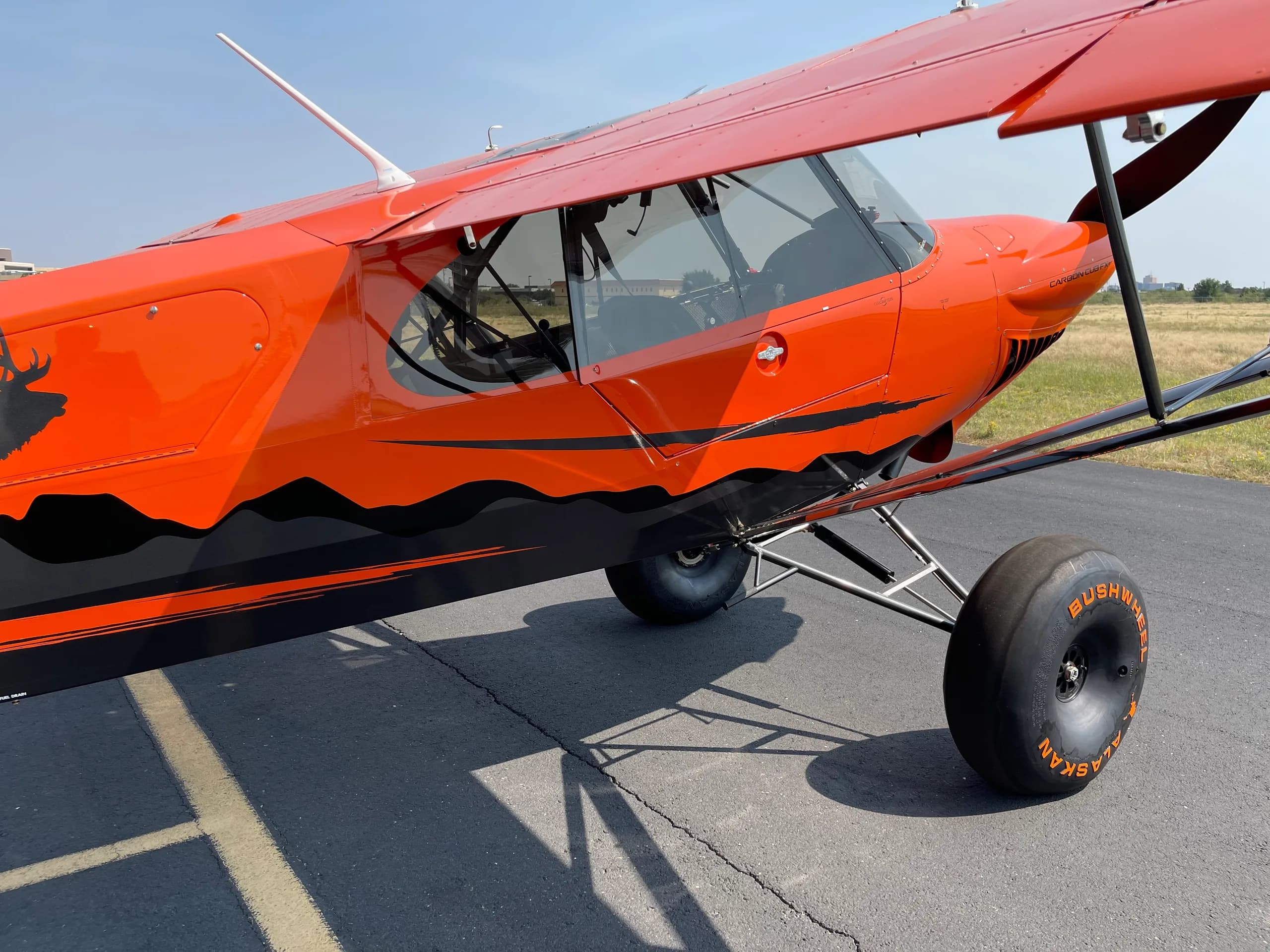 A three-quarter front view of the orange and black Carbon Cub FX-3 aircraft, showing the cockpit and large bush wheels on an airport tarmac.