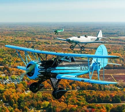 Three biplanes fly in formation over a forest in autumn.