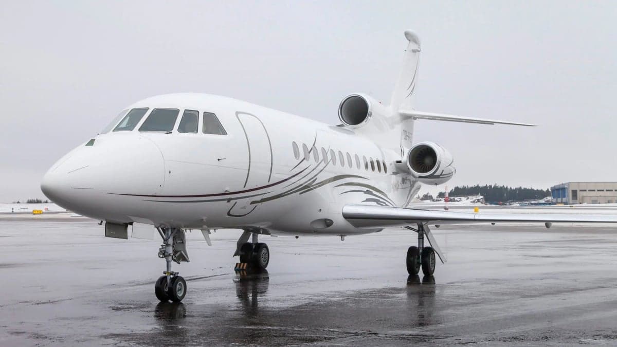 A white Dassault Falcon 900 business jet with grey stripes parked on a wet tarmac in a winter setting.