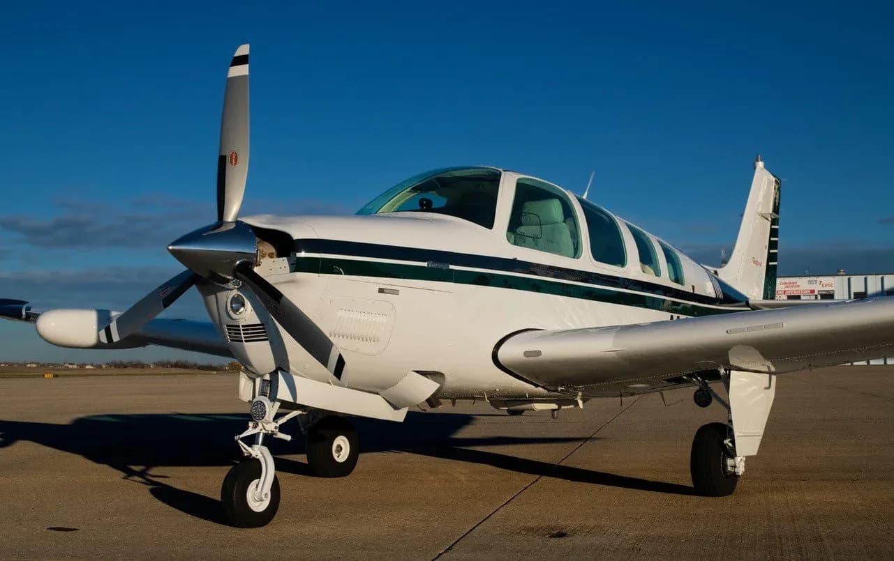A white propeller aircraft with a dark green stripe, possibly a Beechcraft Baron, parked on an airfield under a clear blue sky.