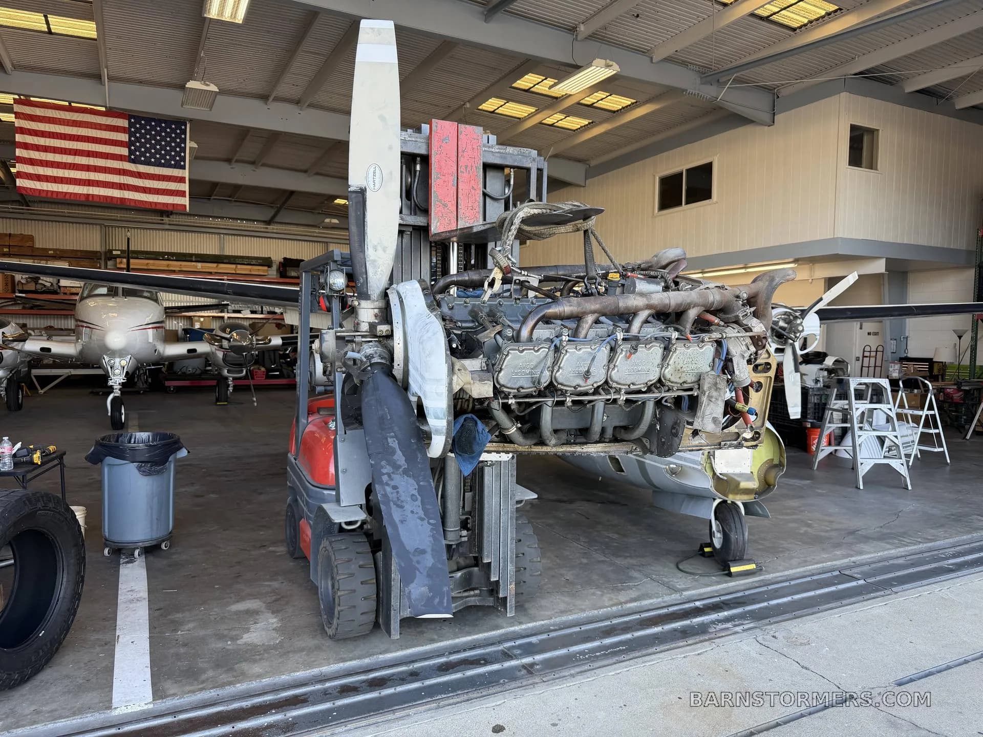 A large aircraft engine, exposed and mounted on a forklift, being worked on inside a hangar with an American flag in the background.