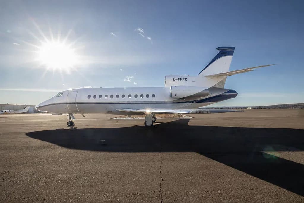 A white and blue Dassault Falcon 900EX business jet with registration C-FPFS parked on an airfield with the sun shining brightly.