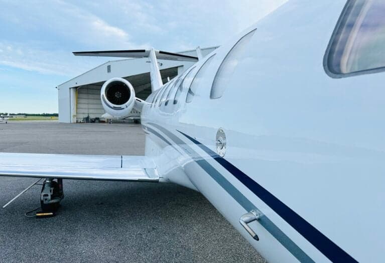 A side view of the fuselage and wing of a white Cessna Citation CJ3 with blue and grey stripes, parked on an asphalt tarmac in front of a hangar.