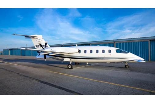 A white Piaggio P.180 Avanti aircraft with black and grey stripes on the tail, parked on an asphalt tarmac in front of blue hangars under a blue sky.
