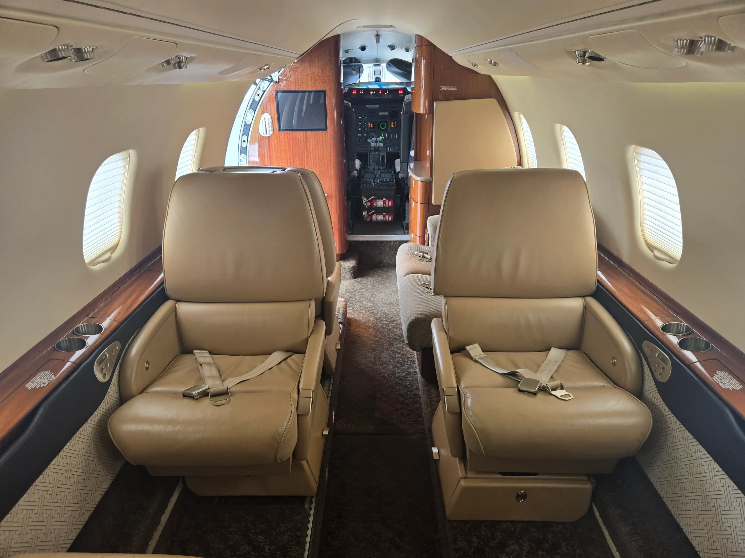 The interior cabin of a business jet, showing two rows of tan leather seats facing each other, with the cockpit visible in the background.
