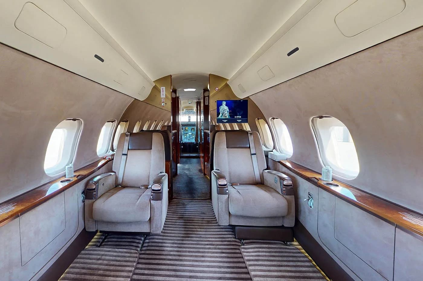 The luxurious interior of a private jet cabin, showing two beige leather seats facing forward with a screen visible in the background.