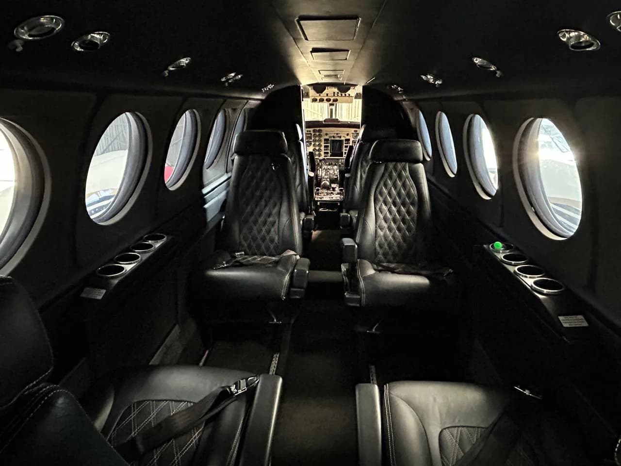 The dark interior cabin of a private aircraft features quilted black leather seats and round windows, looking towards the cockpit.